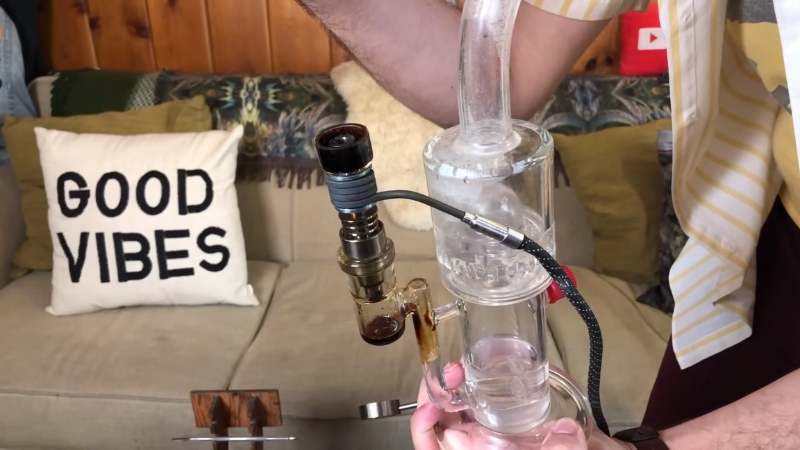 Person holding a dab rig with an electronic nail setup in a living room, preparing to take a dab